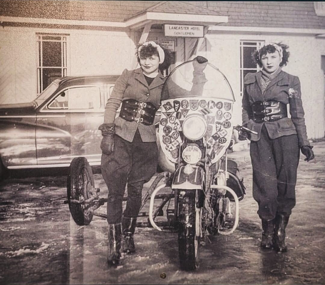 A historic black‑and‑white photograph of two uniformed men standing beside a motorcycle with a sidecar in front of the Lancaster Hotel, capturing early‑20th‑century local law enforcement; this original print hangs inside The Lancaster Smokehouse as part of its heritage display.
